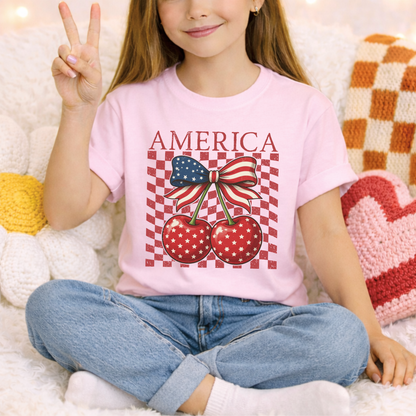 Blossom pink youth America Cherry Bow shirt designed on a Comfort Colors heavyweight tee. Front graphic features patriotic cherries with white stars, green stems, and a red white and blue ribbon bow. Distressed checkerboard detail adds retro boutique style. Soft cotton shirt for girls that pairs easily with denim shorts or sneakers. Perfect for patriotic holidays, summer photos, family trips, and matching sibling or mommy outfits.