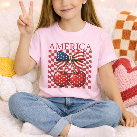 Blossom pink youth America Cherry Bow shirt designed on a Comfort Colors heavyweight tee. Front graphic features patriotic cherries with white stars, green stems, and a red white and blue ribbon bow. Distressed checkerboard detail adds retro boutique style. Soft cotton shirt for girls that pairs easily with denim shorts or sneakers. Perfect for patriotic holidays, summer photos, family trips, and matching sibling or mommy outfits.