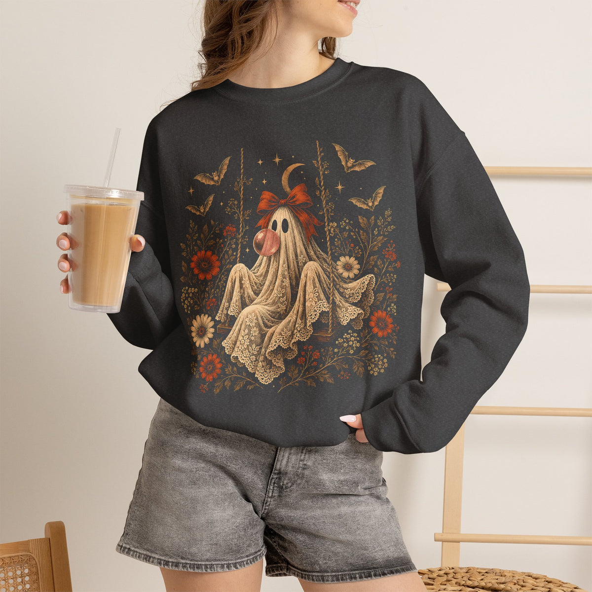 A woman wearing a dark charcoal gray crewneck sweatshirt with a detailed graphic of a ghost on a swing surrounded by autumn leaves and flowers. Vintage Halloween graphic sweater for women featuring a whimsical ghost sitting on a swing while blowing a bubble with bubble gum, surrounded by vintage flowers, botanical elements, bats, lace, stars and a crescent moon in a detailed autumn inspired design.