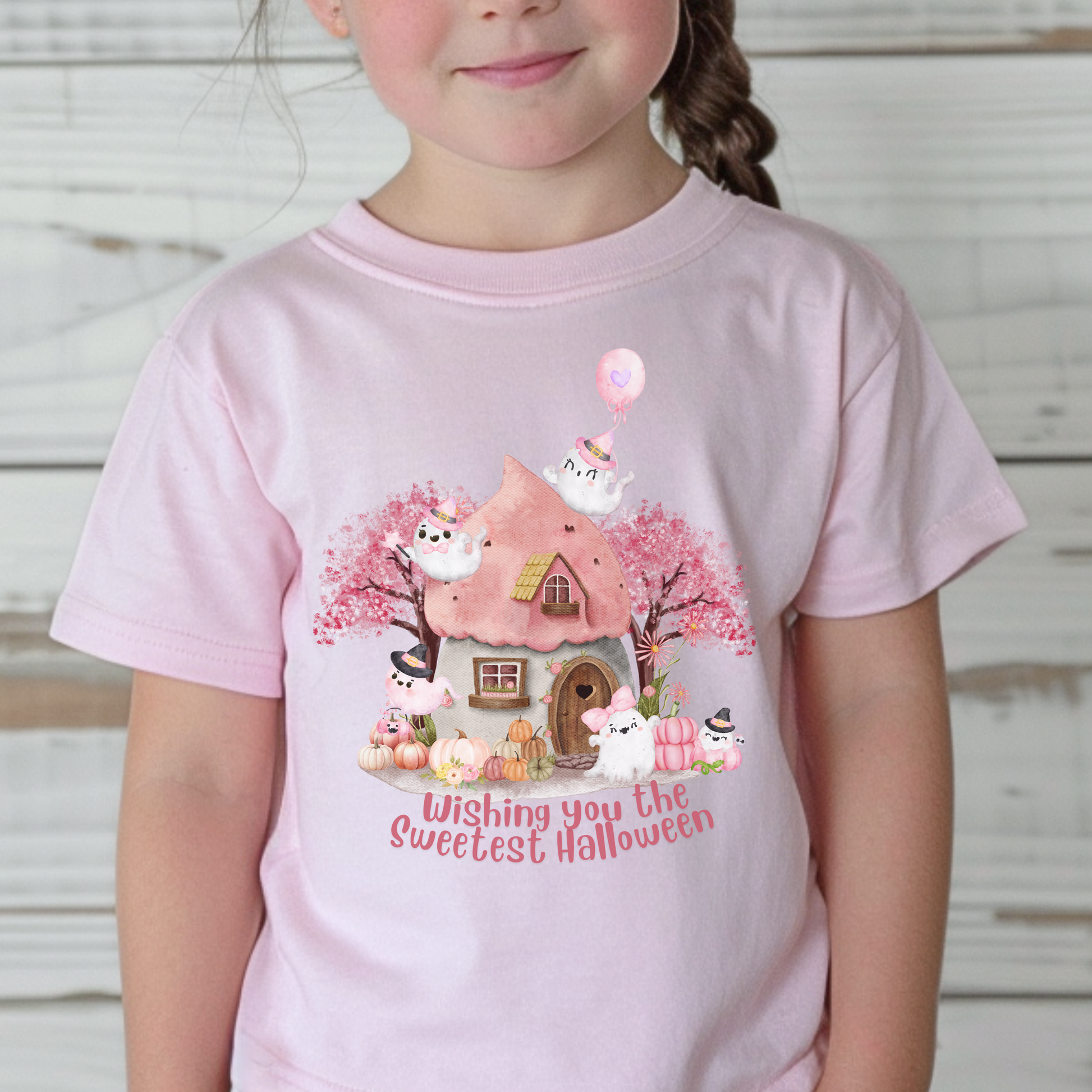 Whimsical pink Halloween shirt with friendly white ghosts, pastel pumpkins, flowers, trees, and a cozy little haunted cottage. The design reads Wishing You the Sweetest Halloween and has a sweet watercolor aesthetic perfect for a toddler Halloween outfit.