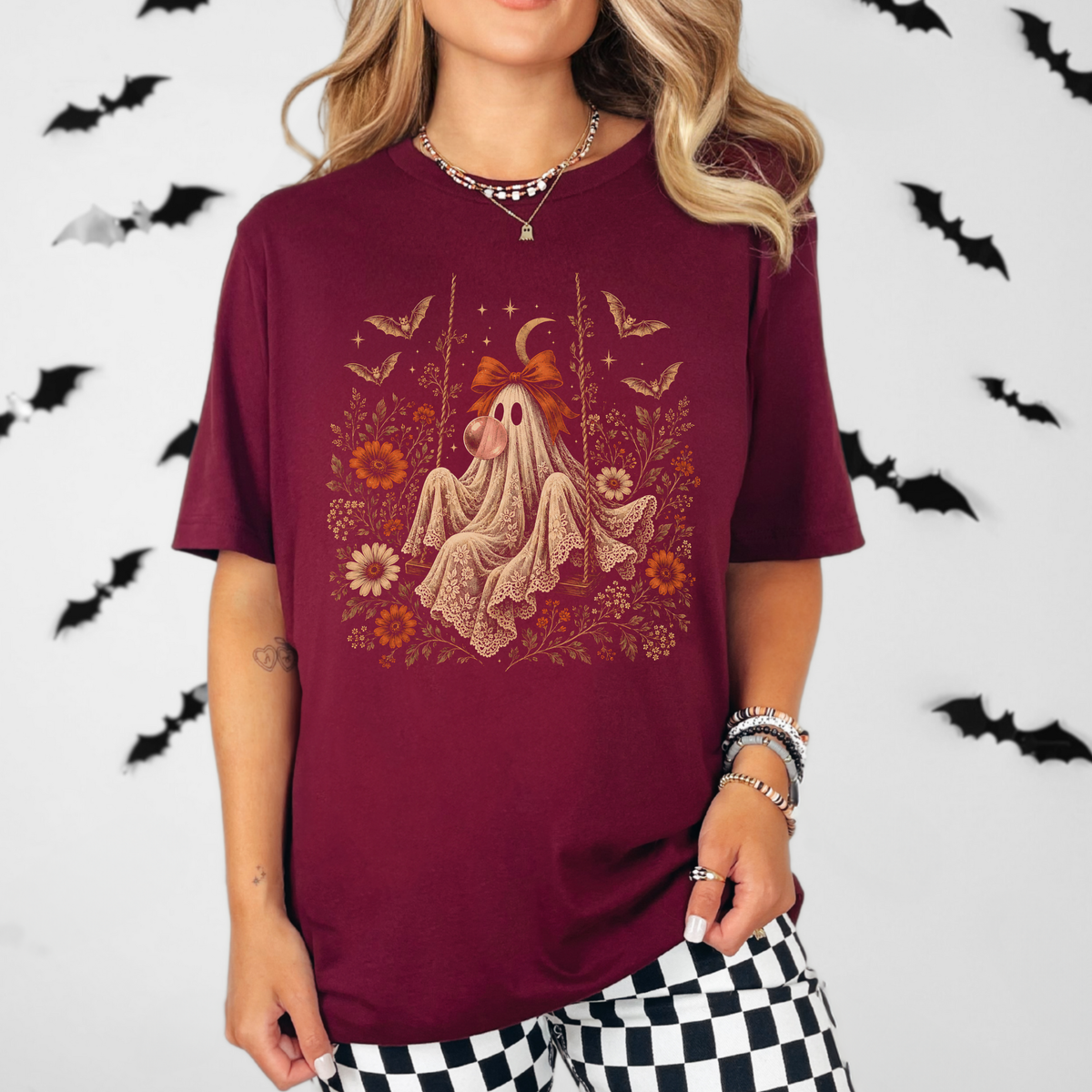 Whimsigoth inspired women's Halloween shirt featuring a vintage ghost seated on a swing and blowing bubble gum. Floral details, botanical foliage, bats, lace, stars and a crescent moon create a romantic spooky season look with vintage gothic character.