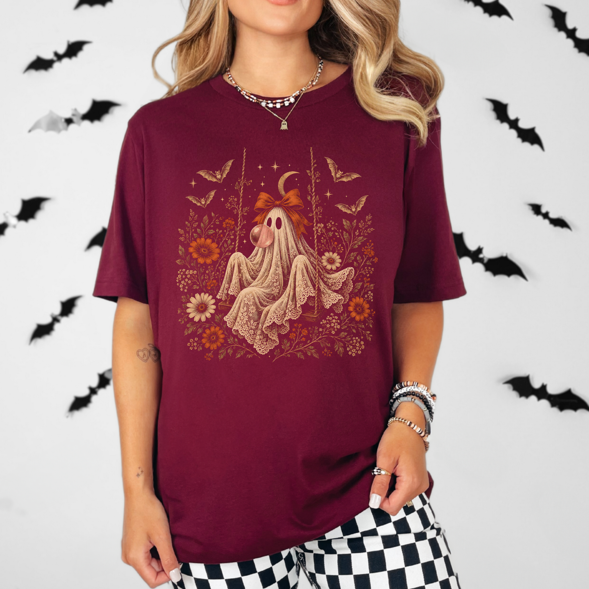 Whimsigoth inspired women's Halloween shirt featuring a vintage ghost seated on a swing and blowing bubble gum. Floral details, botanical foliage, bats, lace, stars and a crescent moon create a romantic spooky season look with vintage gothic character.