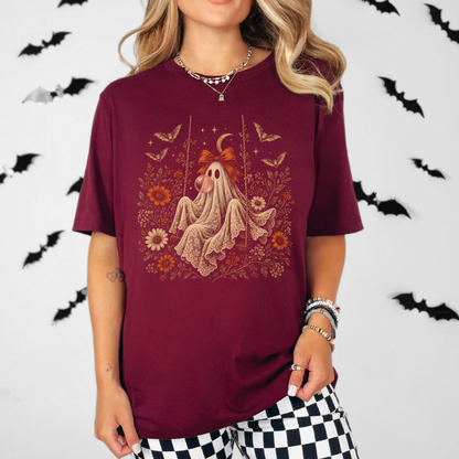 Whimsigoth inspired women's Halloween shirt featuring a vintage ghost seated on a swing and blowing bubble gum. Floral details, botanical foliage, bats, lace, stars and a crescent moon create a romantic spooky season look with vintage gothic character.