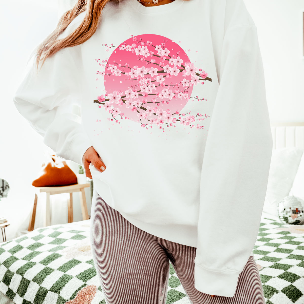 White Comfort Colors 1466 sweatshirt decorated with delicate pink sakura branches and cherry blossoms layered over a soft pink moon. The clean white background lets the floral artwork stand out while creating a feminine, artistic look that pairs easily with leggings, jeans, denim, and casual everyday outfits.