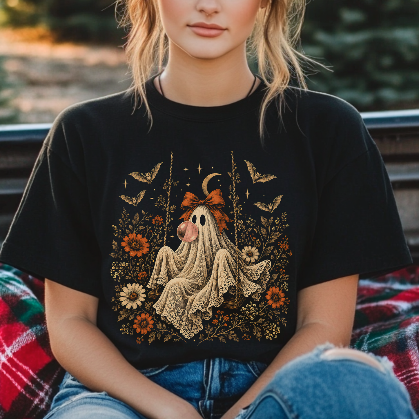 Women's vintage Halloween graphic tee featuring a playful ghost on a swing blowing bubble gum. Floral and botanical elements surround the ghost along with bats, lace, stars and a crescent moon for a whimsical spooky fall aesthetic.