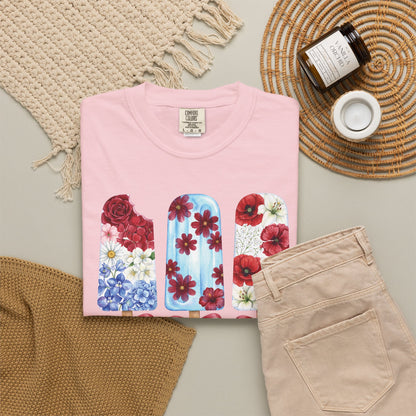 Women's Fourth of July floral T shirt with patriotic popsicle artwork and optional personalization, perfect for Mommy and Me outfits, family reunions and summer parties. A folded light pink t-shirt with a graphic of three floral popsicle designs, styled in a flat lay with beige pants, a woven blanket, a candle jar, and a small cup on a neutral background.