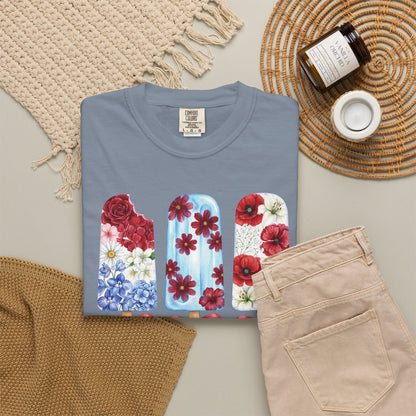 Women's patriotic summer T shirt with watercolor style floral popsicles in red white and blue, a feminine Americana outfit for parades, picnics and cookouts. A folded slate blue t-shirt with a colorful floral popsicle graphic, styled in a flat lay with beige pants, a woven blanket, a candle jar, and a small cup on a neutral background.