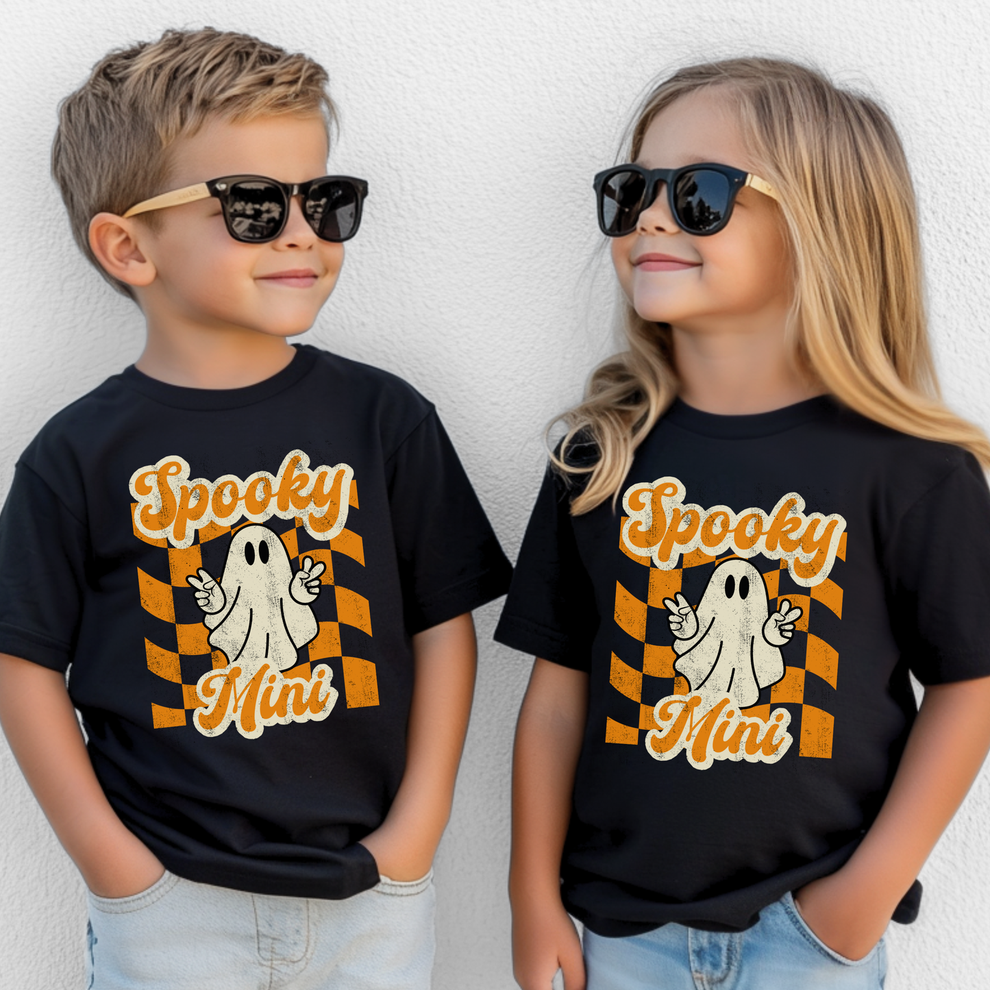 Youth Halloween graphic tee featuring Spooky Mini design with cute ghost peace sign and retro orange checkered background printed on black Bella Canvas 3001Y shirt. Perfect for kids Halloween outfits, youth fall wardrobe, pumpkin patch outings, classroom parties, trick or treating, and spooky season photos. Part of matching family set with toddler Spooky Mini tee and adult Spooky Mama shirt for coordinated Halloween family outfits.