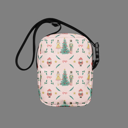 Image: Pink Coquette Nutcracker Utility Crossbody bag. The front of this small mini tote has a Christmas tree, Nutcracker, Sugarplum Fairy, Rocking Horse and a holiday present The rest of the purse is covered in a pattern of ribbons, garland, and Nutcrackers. May be worn across the waist/ hips, across the chest or as a mini backpack, by jaecrece