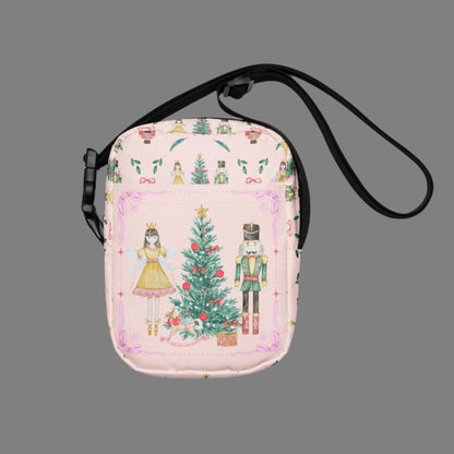 Image: Pink Coquette Nutcracker Utility Crossbody bag. The front of this small mini tote has a Christmas tree, Nutcracker, Sugarplum Fairy, Rocking Horse and a holiday present The rest of the purse is covered in a pattern of ribbons, garland, and Nutcrackers. May be worn across the waist/ hips, across the chest or as a mini backpack, by jaecrece