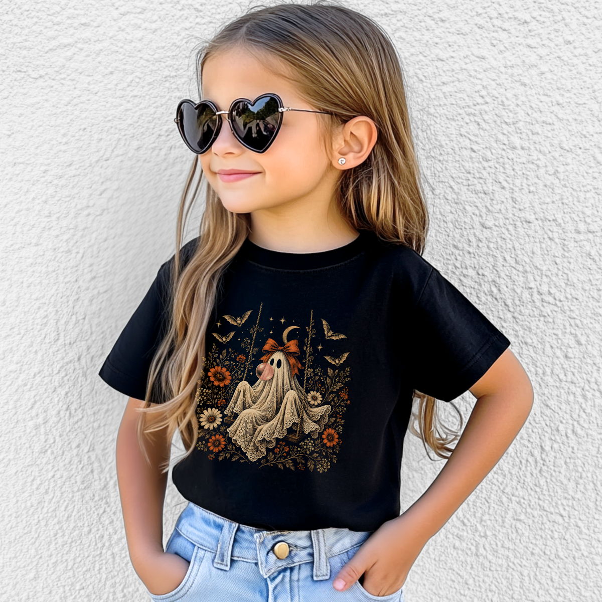 A young girl with long light brown hair wearing heart-shaped sunglasses and a black graphic t-shirt with a ghost illustration, paired with light blue jeans, posing against a white textured wall.