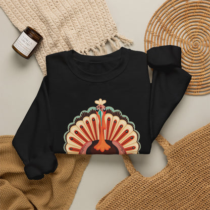 A black long-sleeve crewneck sweatshirt with a colorful peacock graphic on the front, styled in a flat lay with a beige knit blanket, a woven placemat, a straw bag, and a small candle. Colorful cartoon turkey wearing a playful heart expression on a Thanksgiving sweatshirt, perfect for family matching outfits, holiday gatherings and festive fall celebrations