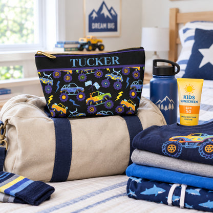 Monster truck toiletry bag packed with a child's travel essentials for summer camp, shown with a duffel bag, folded clothing and other simple belongings. The personalized black zipper pouch features colorful monster trucks, oversized wheels, lightning bolts and racing flags, making it a fun boys travel bag for camp, sleepovers, vacations and family road trips.