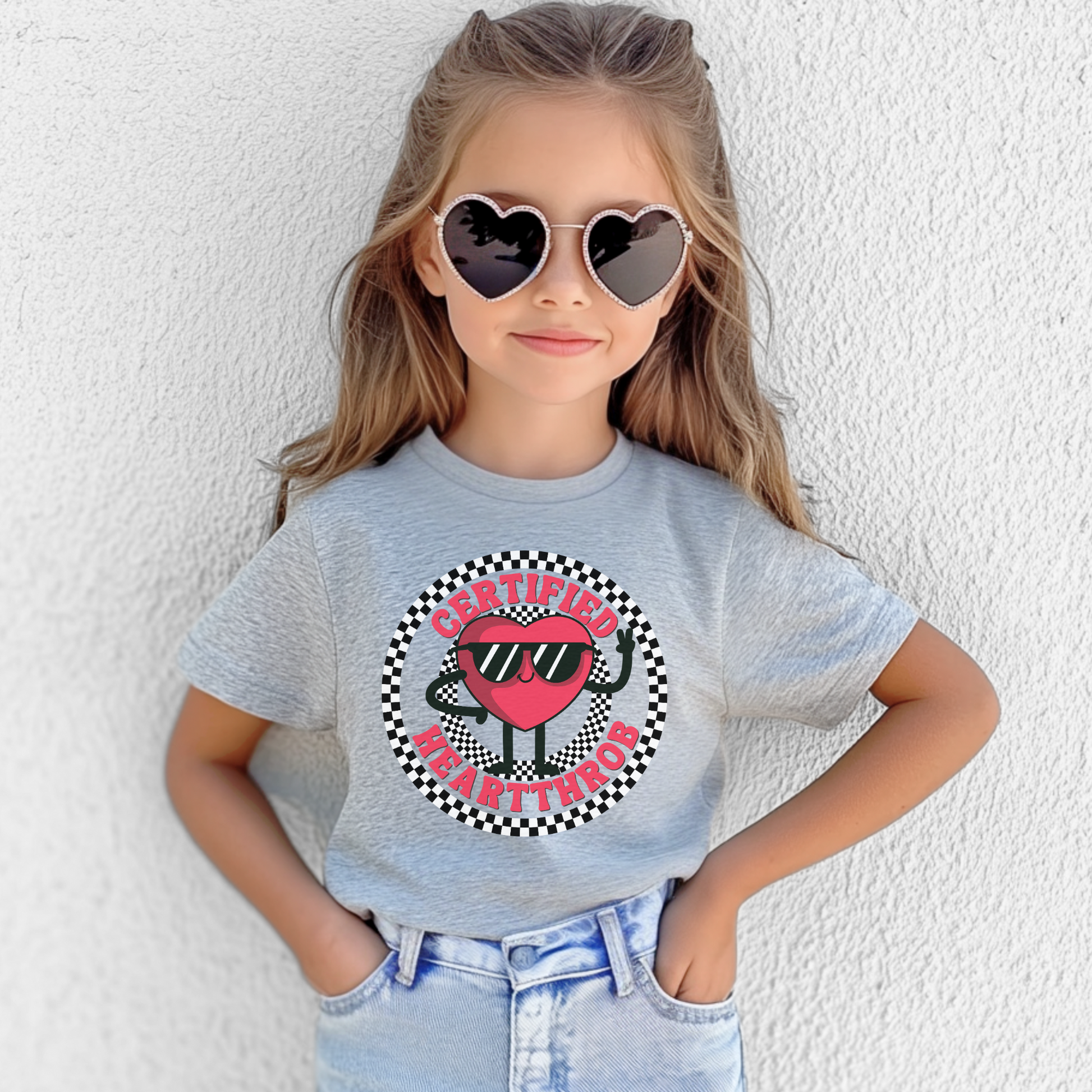 A playful Valentine's Day youth shirt features a cartoon red heart character wearing sunglasses, surrounded by a circular black and white checkerboard design. The words Certified Heartthrob appear around the graphic in bold lettering. Shown on a young girl, the white tee has a fun retro inspired look perfect for holiday celebrations and casual outfits.