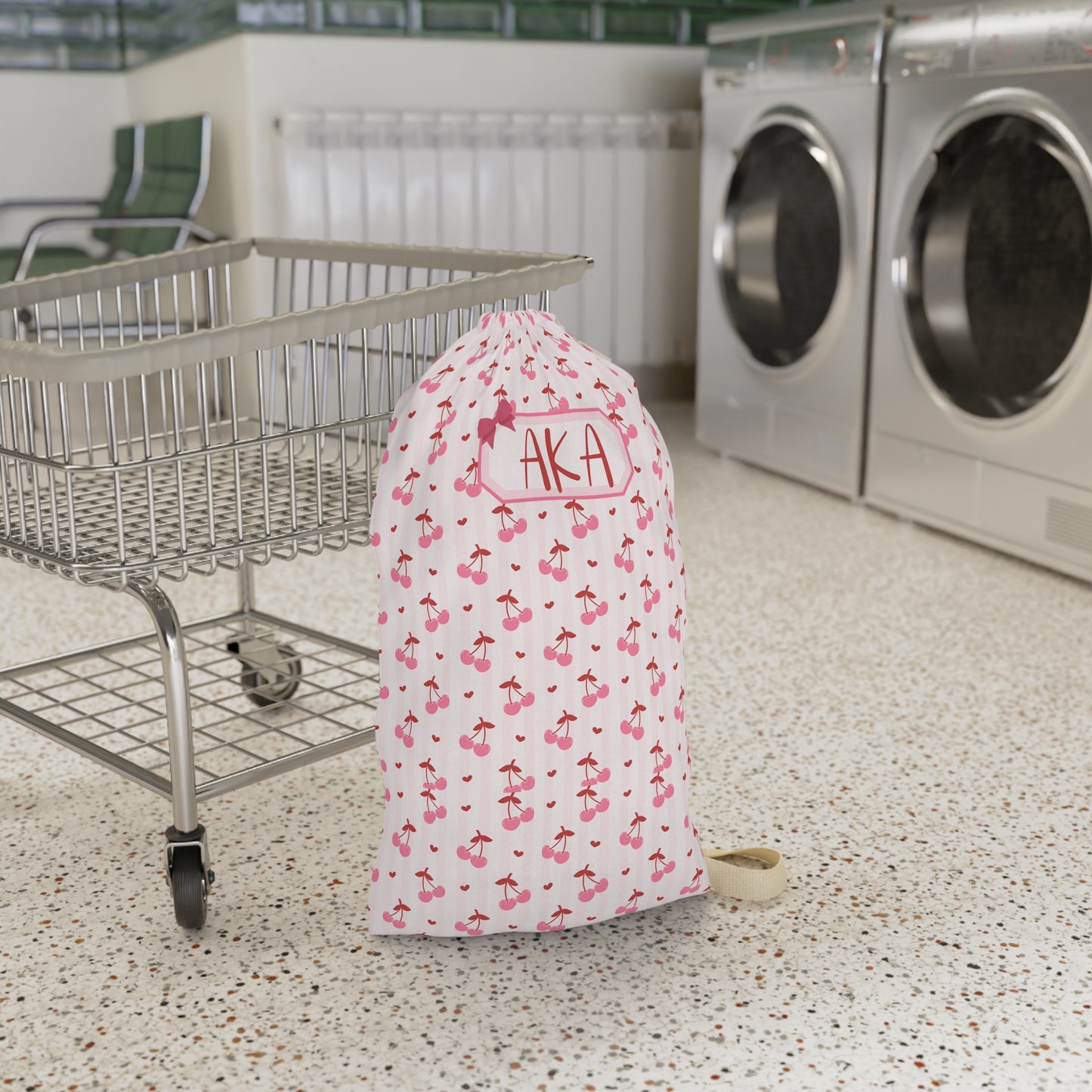 Personalized coquette cherry laundry bag displayed in a laundromat setting, showcasing the custom name personalization and charming cherry pattern. The spacious drawstring design offers plenty of room for dirty clothes, towels, and linens, making it ideal for college dorms, camp, travel, and family vacations. This custom laundry sack combines practical organization with a soft feminine aesthetic and makes a thoughtful gift for students, teens, bridesmaids, and travelers.