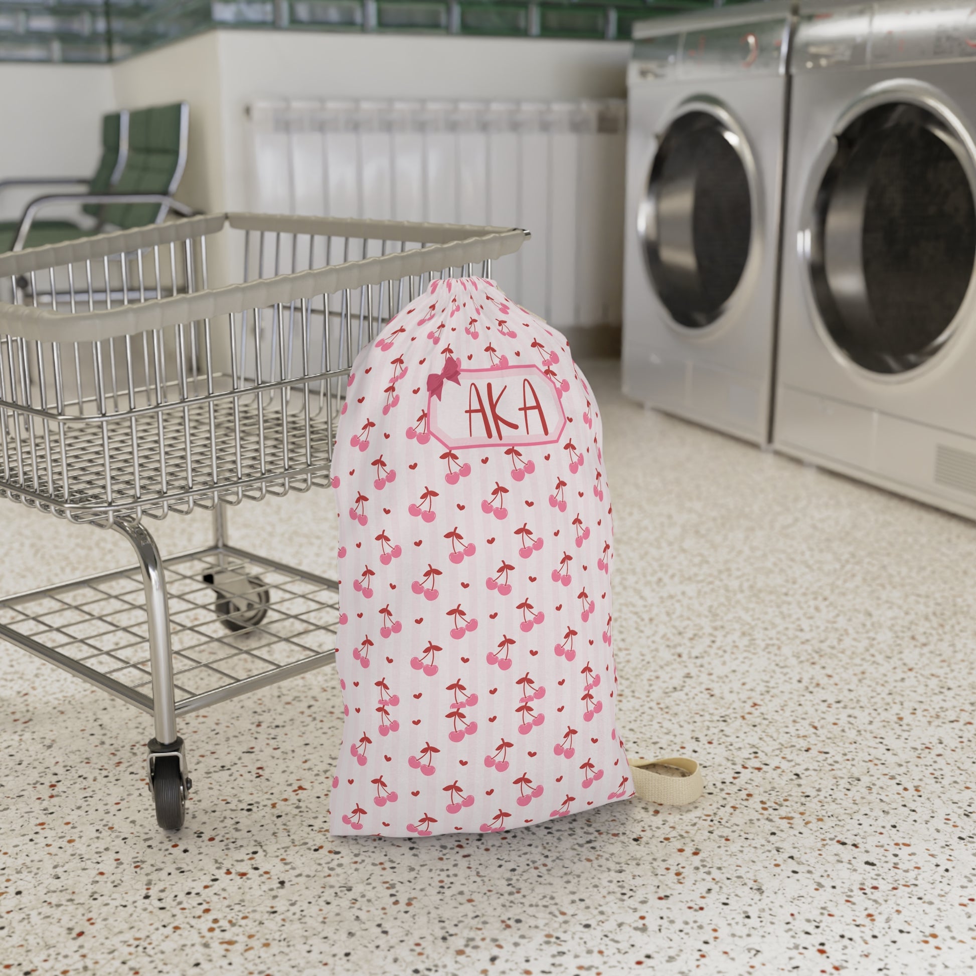 Personalized coquette cherry laundry bag displayed in a laundromat setting, showcasing the custom name personalization and charming cherry pattern. The spacious drawstring design offers plenty of room for dirty clothes, towels, and linens, making it ideal for college dorms, camp, travel, and family vacations. This custom laundry sack combines practical organization with a soft feminine aesthetic and makes a thoughtful gift for students, teens, bridesmaids, and travelers.