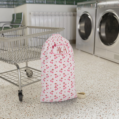Personalized coquette cherry laundry bag displayed in a laundromat setting, showcasing the custom name personalization and charming cherry pattern. The spacious drawstring design offers plenty of room for dirty clothes, towels, and linens, making it ideal for college dorms, camp, travel, and family vacations. This custom laundry sack combines practical organization with a soft feminine aesthetic and makes a thoughtful gift for students, teens, bridesmaids, and travelers.