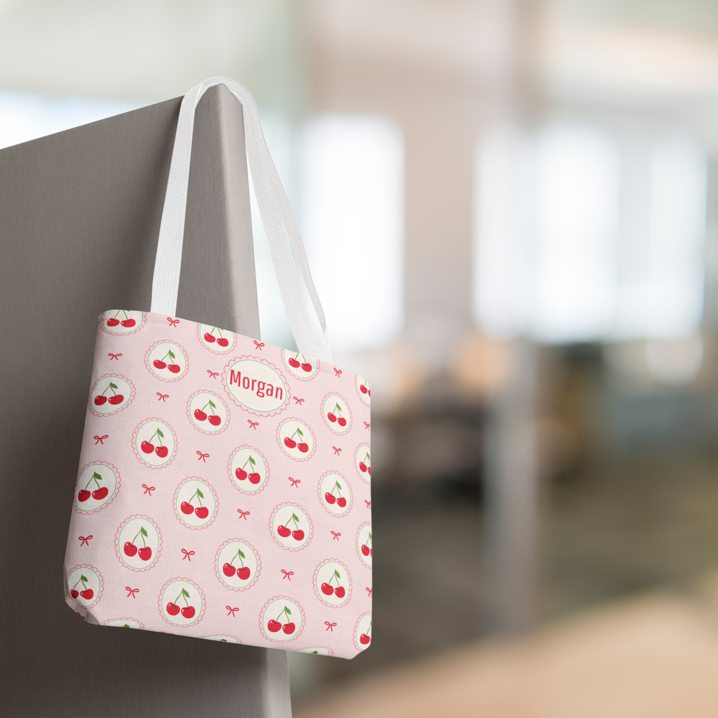 A pink canvas tote bag with a cherry print pattern and white straps, hanging on a gray pillar in a blurred indoor setting. The bag has a circular patch that reads 'Morgan'. Custom cherry tote bag in a soft pink palette featuring repeating cherries and charming bow accents. Personalized with a name or monogram, this pastel fruit pattern bag is a cute Cutecore and Kawaii accessory for tweens, teens, teachers, back to school, summer vacations, beach days, shopping, or everyday use.