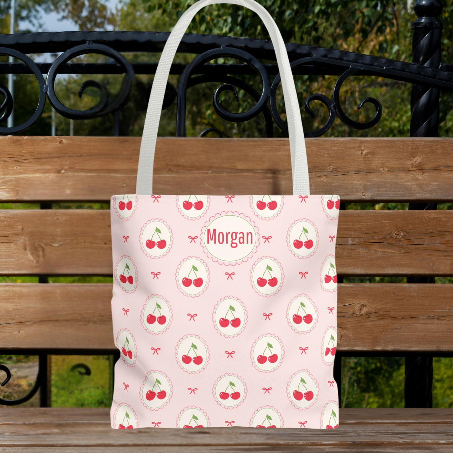 A light pink tote bag with a repeating cherry print and a personalized name patch reading 'Morgan', displayed on a wooden bench outdoors. Custom pink cherry bag with a feminine bow pattern and pastel fruit design. Personalize this cute everyday bag with your name or monogram. The Cutecore, Kawaii, and Coquette inspired aesthetic makes it a fun accessory for teenagers, tweens, teachers, shopping, groceries, beach trips, and vacations.