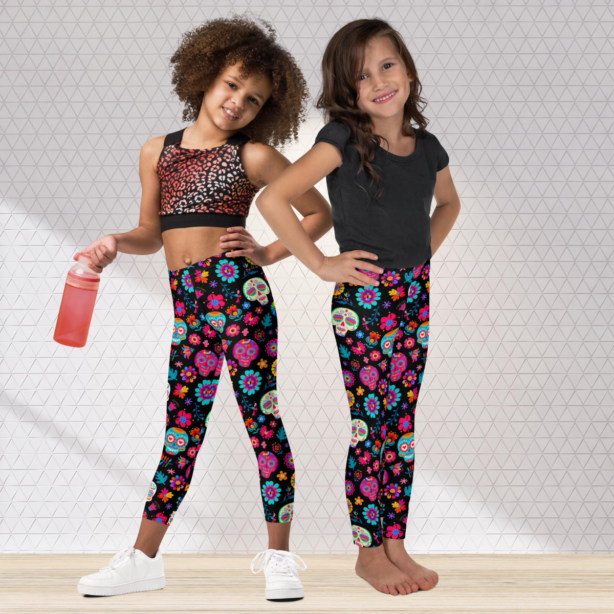 Two tween girls standing side by side wearing matching Día de los Muertos leggings with colorful sugar skulls, flowers, and festive details on a black background. The high waist, full length leggings create a coordinated look for Halloween, Day of the Dead celebrations, or everyday wear, with girls sizes available from 2T through 20.