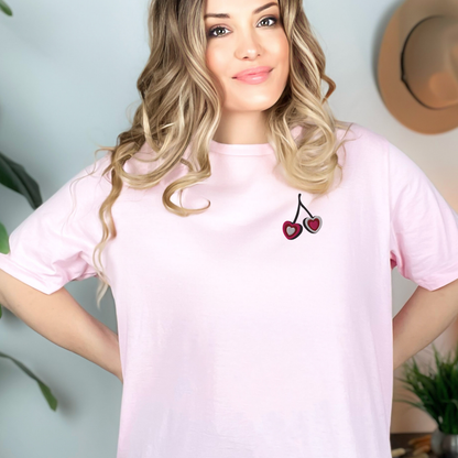 Woman wearing light pink embroidered cherry hearts t shirt with delicate red cherry heart embroidery on the front. Designed on a relaxed fit Gildan S5000 tee, this soft feminine color is perfect for casual wear, weekend outings, and everyday comfort. A matching girls shirt is available separately for mommy and me outfits, making it a sweet coordinated family fashion option.