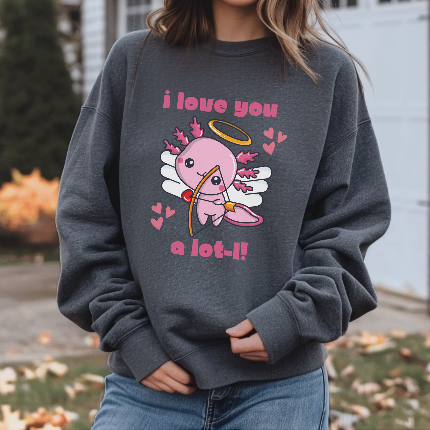 A person wearing a dark gray crewneck sweatshirt with a pink angel graphic and the text 'i love you a lot-!!' on the front, paired with blue jeans in an outdoor setting. Cute axolotl lover sweatshirt with a playful Valentine’s Day cupid design and romantic animal pun. Cozy unisex crewneck perfect for Valentine celebrations, Galentine’s gatherings, animal lover gifts, casual outfits, and coordinated family Valentine photos.