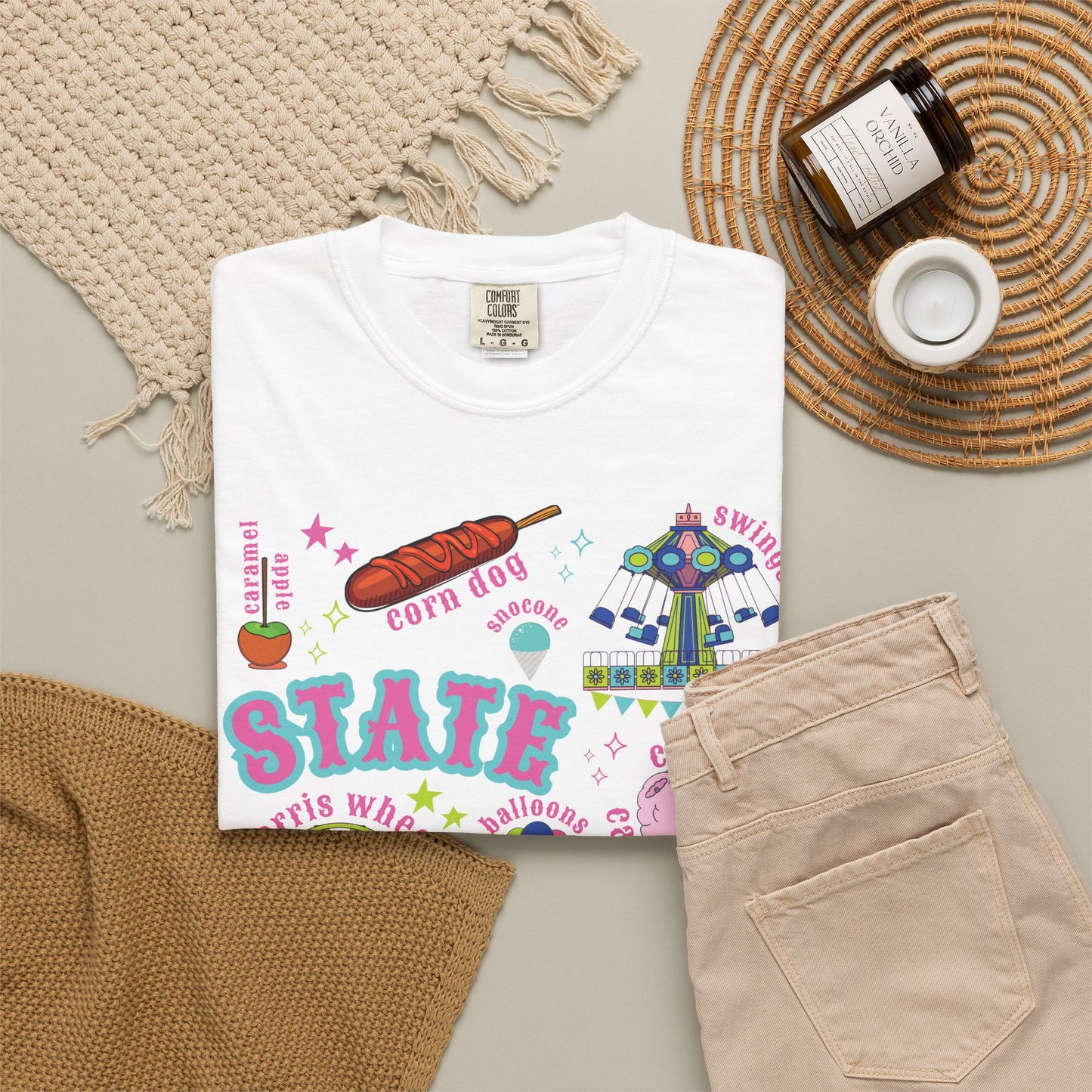A nostalgic summer fair graphic on a T shirt features bold STATE FAIR lettering surrounded by a Ferris wheel, roller coaster, carnival swing ride, cotton candy, corn dogs, funnel cake, balloons, livestock, blue ribbon and ticket illustrations with colorful stars for a colorful fair day. The design blends carnival fun with classic Americana.