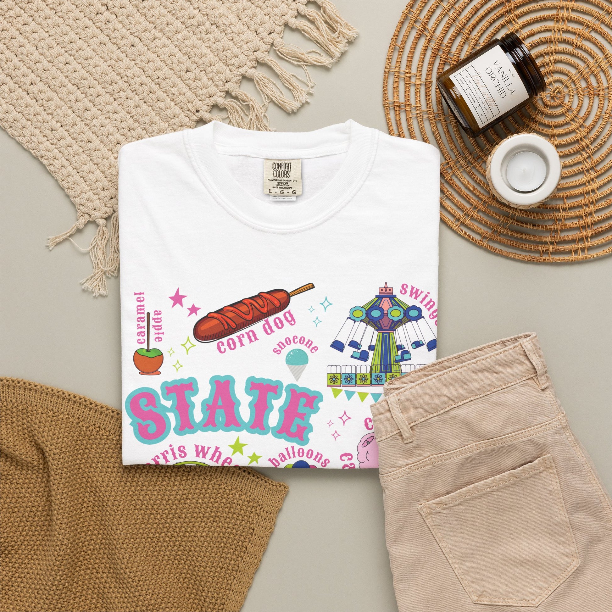A nostalgic summer fair graphic on a T shirt features bold STATE FAIR lettering surrounded by a Ferris wheel, roller coaster, carnival swing ride, cotton candy, corn dogs, funnel cake, balloons, livestock, blue ribbon and ticket illustrations with colorful stars for a colorful fair day. The design blends carnival fun with classic Americana.