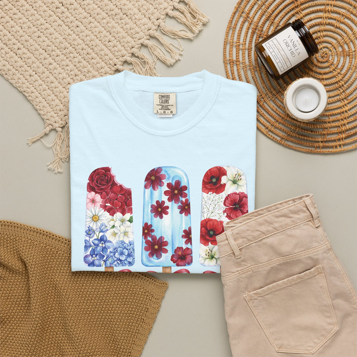 A folded light blue T-shirt with a colorful floral popsicle graphic, styled in a flat lay with beige pants, a woven blanket, a candle jar, and a small cup on a neutral background. Women's red white and blue floral T shirt with whimsical popsicle artwork, available in True Navy, Chambray, White, Ivory, Blossom and Blue Jean
