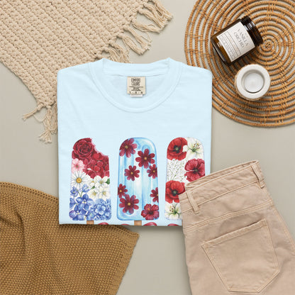A folded light blue T-shirt with a colorful floral popsicle graphic, styled in a flat lay with beige pants, a woven blanket, a candle jar, and a small cup on a neutral background. Women's red white and blue floral T shirt with whimsical popsicle artwork, available in True Navy, Chambray, White, Ivory, Blossom and Blue Jean