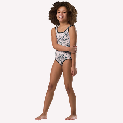 A young girl with curly brown hair stands barefoot against a solid white background, wearing a sleeveless one-piece swimsuit with a navy blue floral toile pattern on a pink base.