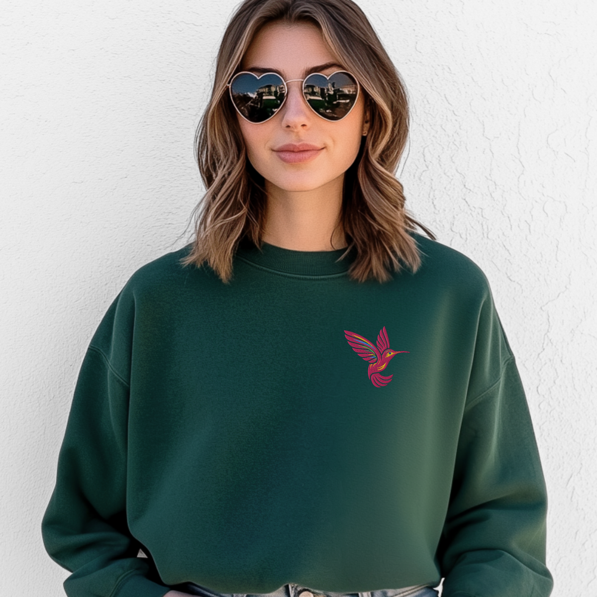 Woman wearing a forest green Gildan crewneck sweatshirt featuring a colorful embroidered hummingbird on the chest. The relaxed unisex sweatshirt has a comfortable everyday fit and the rich green fabric complements the nature inspired bird design. This casual look shows how the embroidered hummingbird sweatshirt can be styled with everyday outfits and is a thoughtful choice for bird lovers, hummingbird enthusiasts, gardeners, and nature lovers.