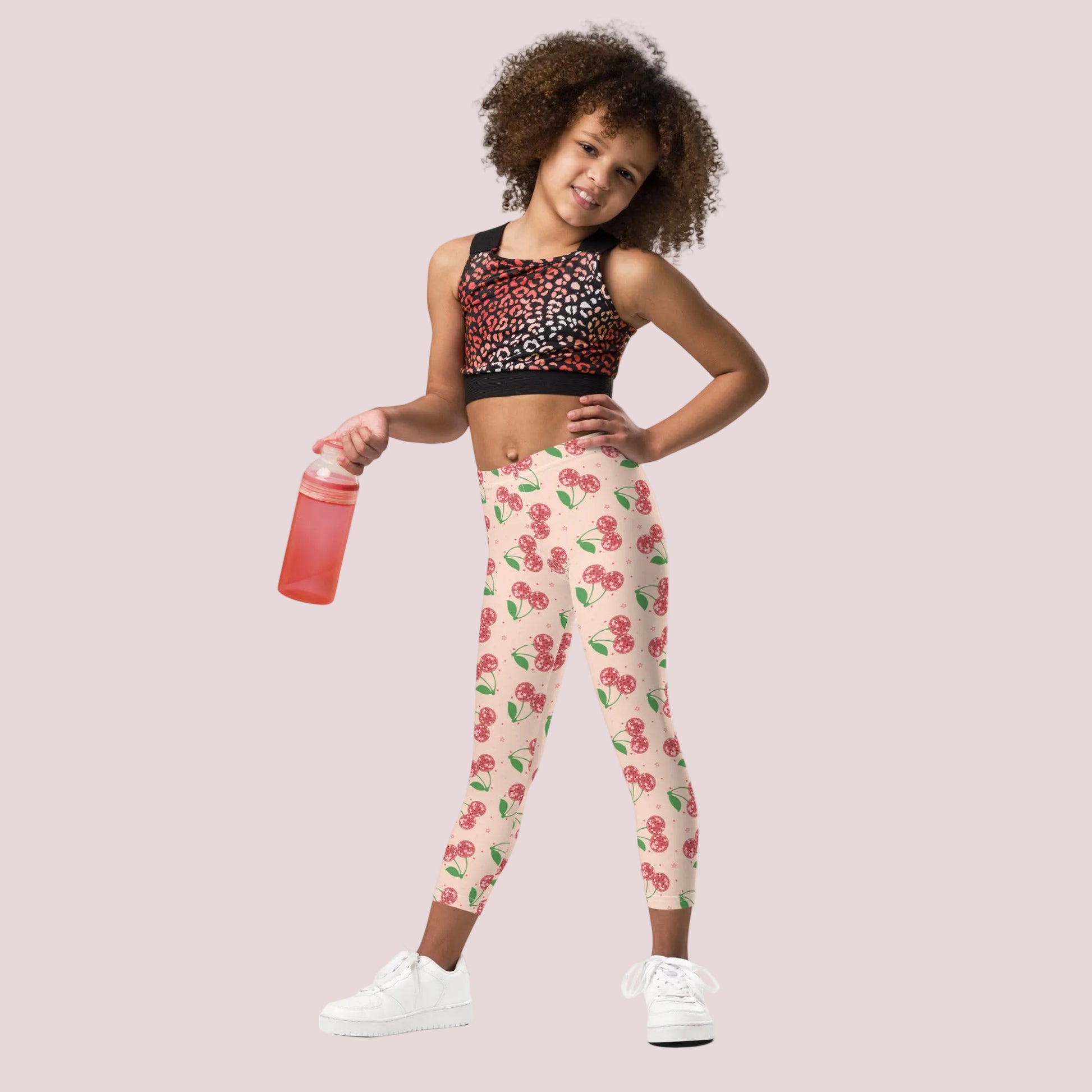 Young girl modeling cherry print leggings with a soft pink background and red cherries, paired with a sporty crop top and white sneakers. She is holding a water bottle and standing in a confident pose, showing the high rise fit and full length design. These leggings are perfect for active days, dance practice, or casual outfits, offering both comfort and playful style for girls who love fun patterns.