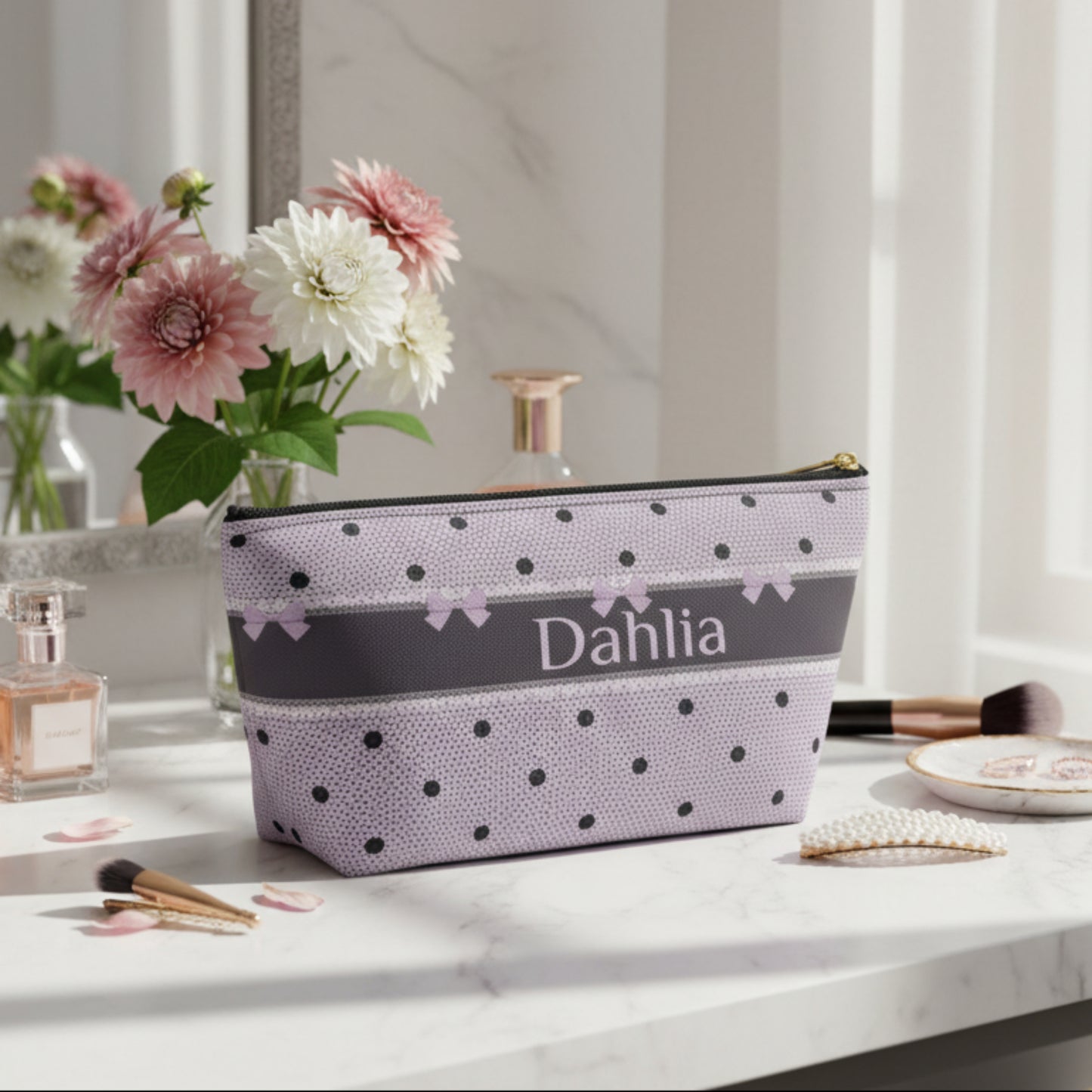 A personalized zippered pouch with a lavender and black polka dot pattern, featuring a dark gray horizontal band with the name 'Dahlia' in white script and small lavender bows. The pouch has a gold zipper pull and is photographed on a clean white background.