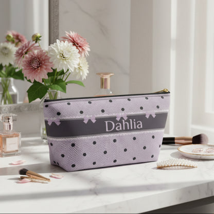 A personalized zippered pouch with a lavender and black polka dot pattern, featuring a dark gray horizontal band with the name 'Dahlia' in white script and small lavender bows. The pouch has a gold zipper pull and is photographed on a clean white background.