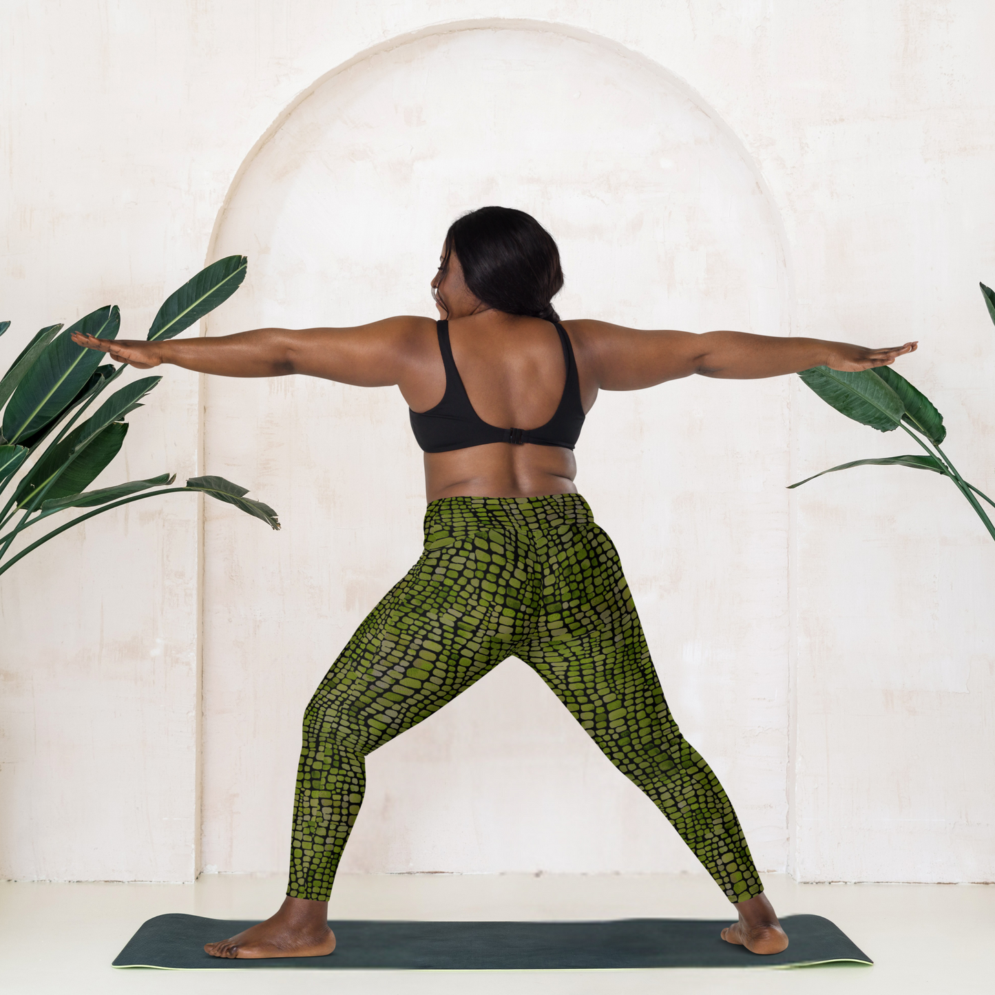 Person practicing yoga in a studio with plants and a white wall. The woman is wearing green reptile print yoga pants 