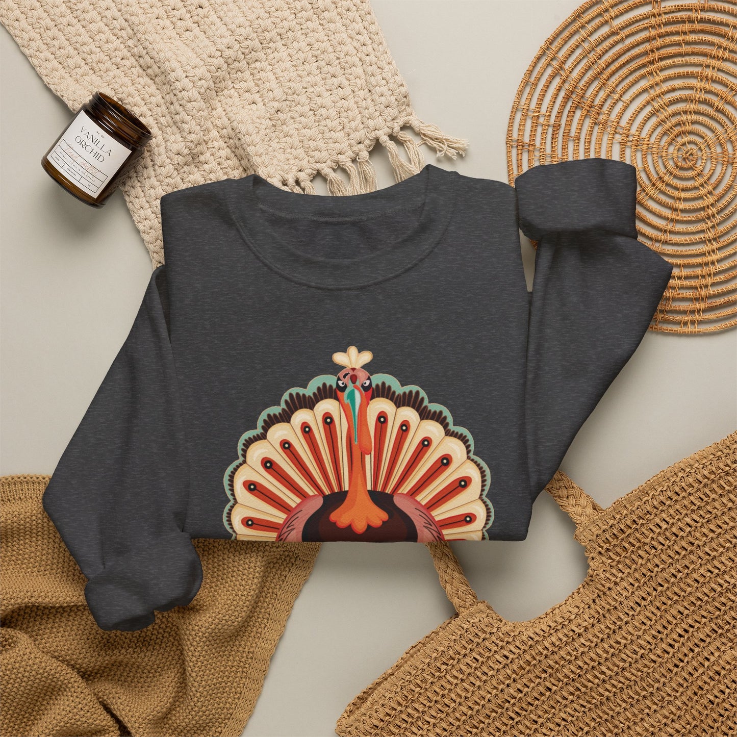 A dark heather gray crewneck sweatshirt with a colorful Thanksgiving turkey graphic on the front, styled in a flat lay with a beige knit blanket, a woven basket, a straw bag, and a small candle. Cute Thanksgiving sweatshirt featuring a colorful turkey creating a heart with its wings, a fun festive fall outfit for moms, dads, teens and kids