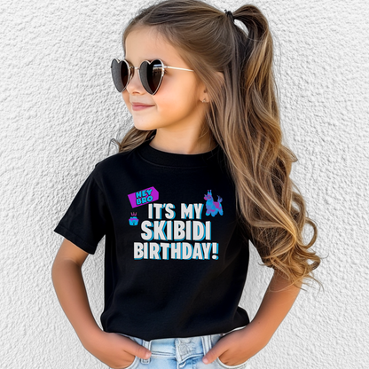 A young girl with long wavy brown hair in a ponytail, wearing heart-shaped sunglasses and a black graphic t-shirt that reads 'IT'S MY SKIBIDI BIRTHDAY!' in white and blue, paired with light blue jeans, standing against a white textured wall. Youth birthday graphic tee featuring the phrase It’s My Skibidi Birthday with colorful playful graphics and bold blue and white lettering, a funny kids birthday shirt for parties and birthday celebrations