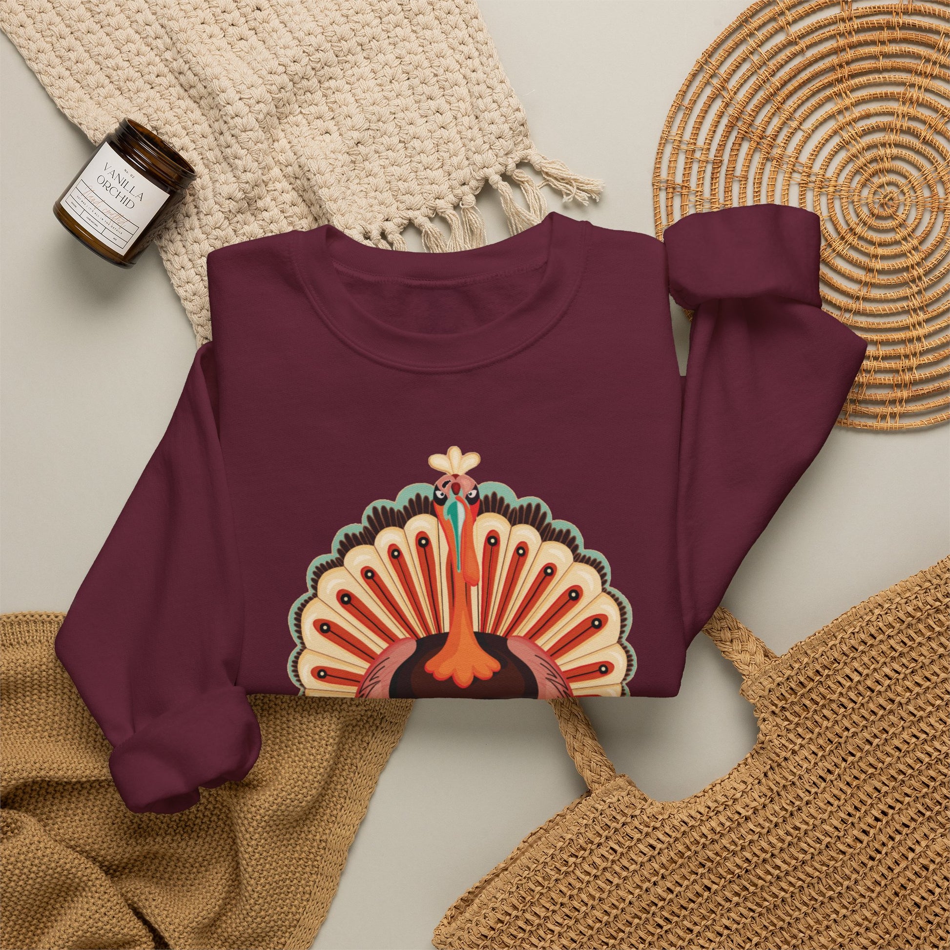 A folded long sleeve crewneck sweatshirt in deep burgundy, featuring a colorful graphic of a peacock with its tail feathers fanned. The sweatshirt is styled in a flat lay with a beige knit blanket, a woven straw bag, a small candle jar, and a round woven placemat on a light gray background.