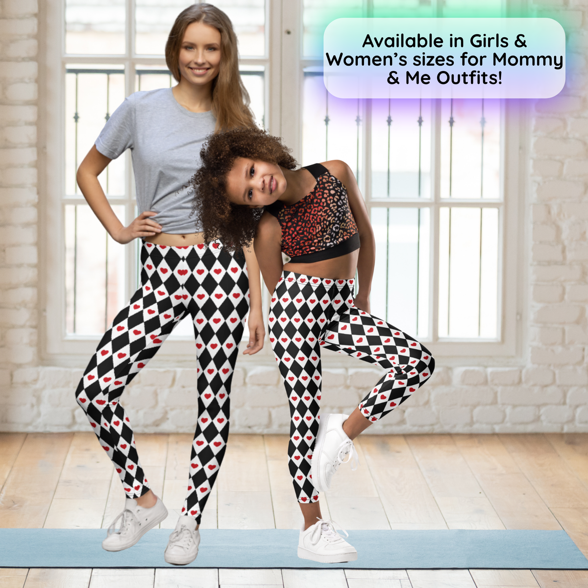 A mother and daughter wear matching black and white harlequin leggings decorated with bright red hearts. The coordinated outfits show how the playful design can be styled as a Mommy and Me look for Valentine's Day, family photos, Halloween, or a fun day together. The girls leggings feature a full length high waisted design, while the matching adult leggings create a cute coordinated look.
