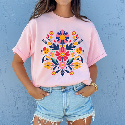Light pink Gildan 64000 Softstyle tee featuring a colorful Otomi inspired floral design with Mexican folk art elements. The soft pink shirt contrasts with the vibrant flowers to create a playful feminine take on a Mexican folk art graphic tee. Great for casual outfits, Hispanic Heritage Month, fiestas, family photos, cultural celebrations, and anyone drawn to colorful floral and folk art inspired clothing.