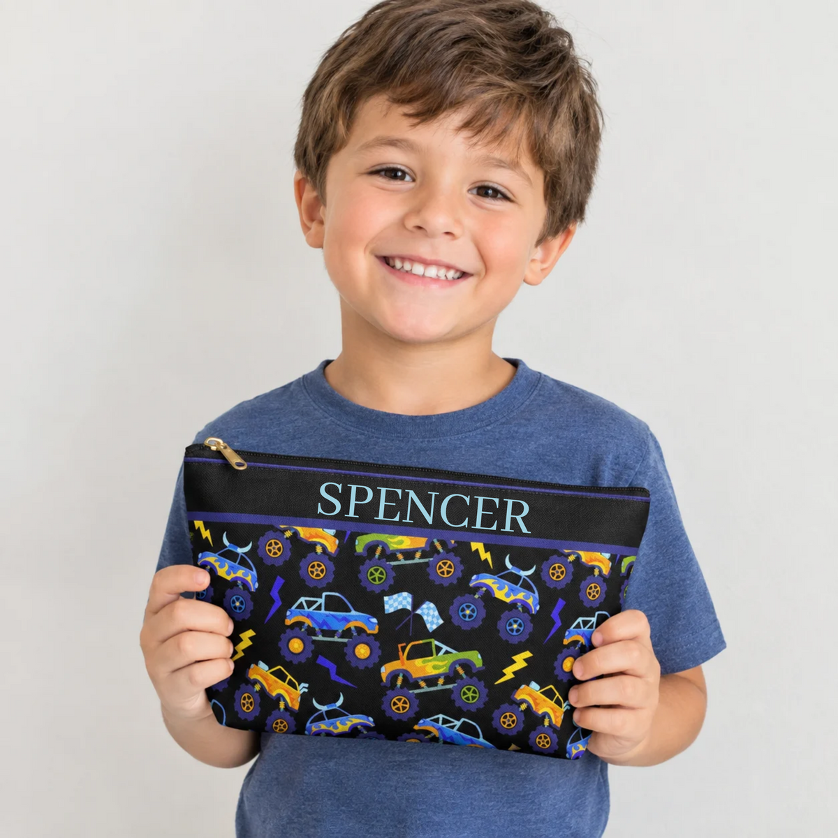 Smiling young boy holding a black personalized kids toiletry bag featuring colorful monster trucks, oversized wheels, lightning bolts and checkered racing flags in blue, purple, yellow, orange and green. The boys travel pouch has a zipper closure and a fun racing inspired design, showing the size and style of the bag for kids, sleepovers, summer camp, vacations and road trips.