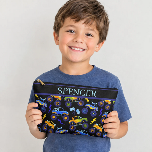 Smiling young boy holding a black personalized kids toiletry bag featuring colorful monster trucks, oversized wheels, lightning bolts and checkered racing flags in blue, purple, yellow, orange and green. The boys travel pouch has a zipper closure and a fun racing inspired design, showing the size and style of the bag for kids, sleepovers, summer camp, vacations and road trips.