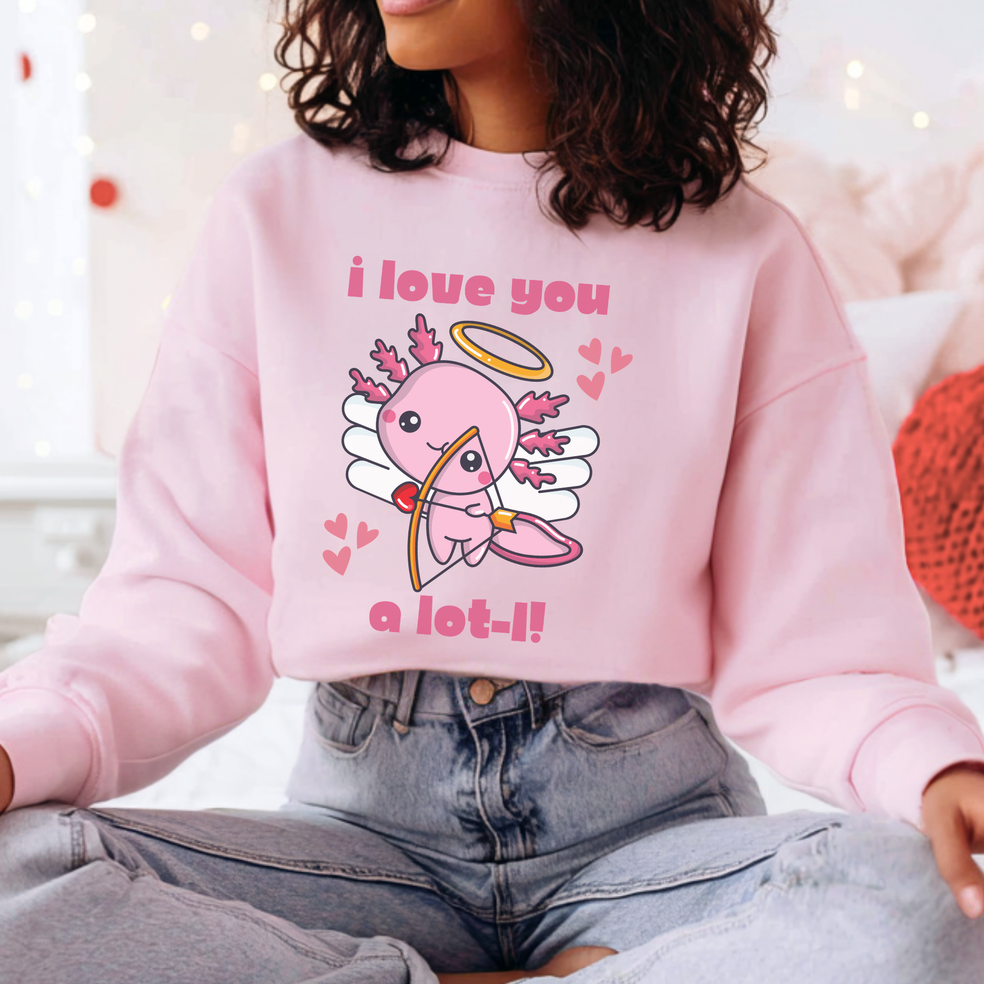 A person wearing a light pink crewneck sweatshirt with a cute cartoon axolotl graphic and the text 'i love you a lot-!!' paired with light-wash jeans in a softly lit indoor setting. Funny axolotl Valentine sweatshirt featuring a cute pink cupid axolotl graphic with an I love you a lotl message. Playful Valentine’s Day clothing for animal lovers, couples, friends, classroom celebrations, family photos, and cozy winter outfits.