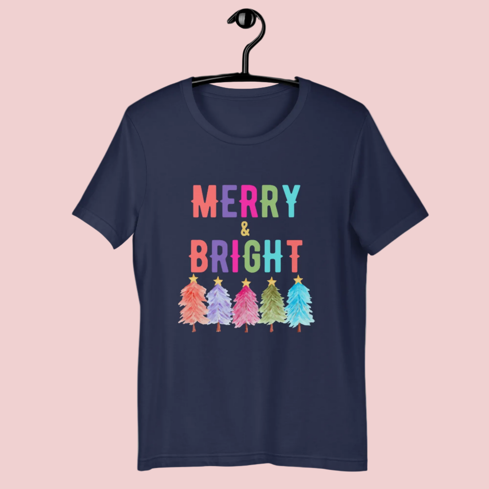 Rainbow Merry and Bright Christmas shirt with colorful trees and bold festive text printed on a Bella Canvas 3001 tee, styled in a cozy holiday home with pink Christmas tree and warm lights, perfect for Christmas parties, family photos, teacher gifts, cute holiday shirts for women, unisex festive clothing, boutique Christmas apparel, cozy winter outfit, trendy graphic tee