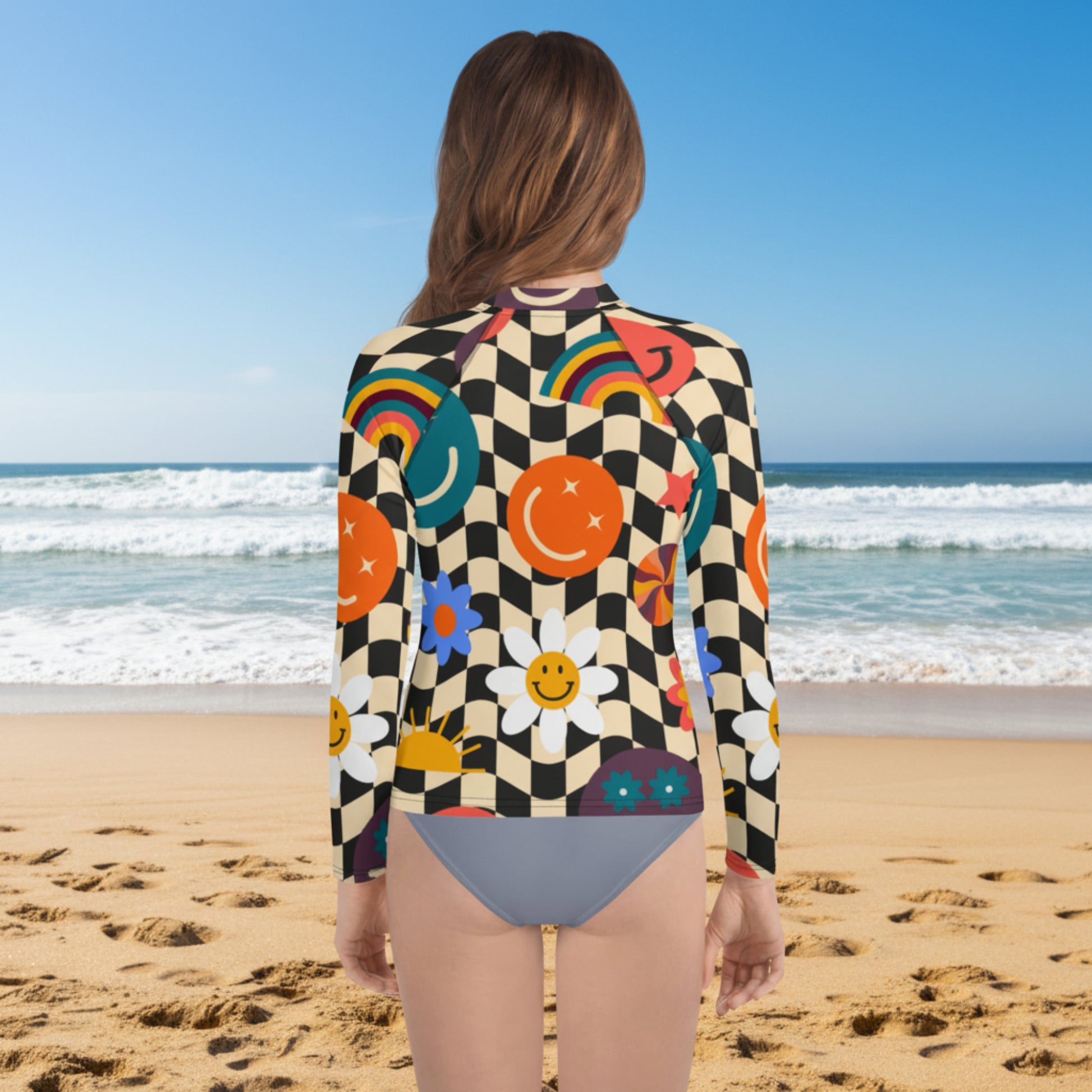 Back view of a teen girl wearing a retro wavy checkerboard long sleeve rash guard on a sandy beach. The beige and black groovy checker print features colorful smiley faces, rainbows, daisies, stars, and swirls. UPF 50+ girls swim shirt available in sizes 2T–20 with coordinating family swimwear, including girls swimsuits, women's swimsuits, and men's swim trunks.