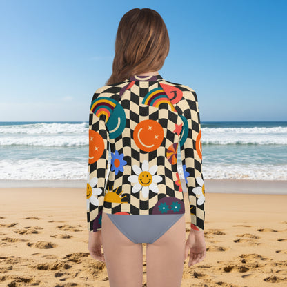 Back view of a teen girl wearing a retro wavy checkerboard long sleeve rash guard on a sandy beach. The beige and black groovy checker print features colorful smiley faces, rainbows, daisies, stars, and swirls. UPF 50+ girls swim shirt available in sizes 2T–20 with coordinating family swimwear, including girls swimsuits, women's swimsuits, and men's swim trunks.