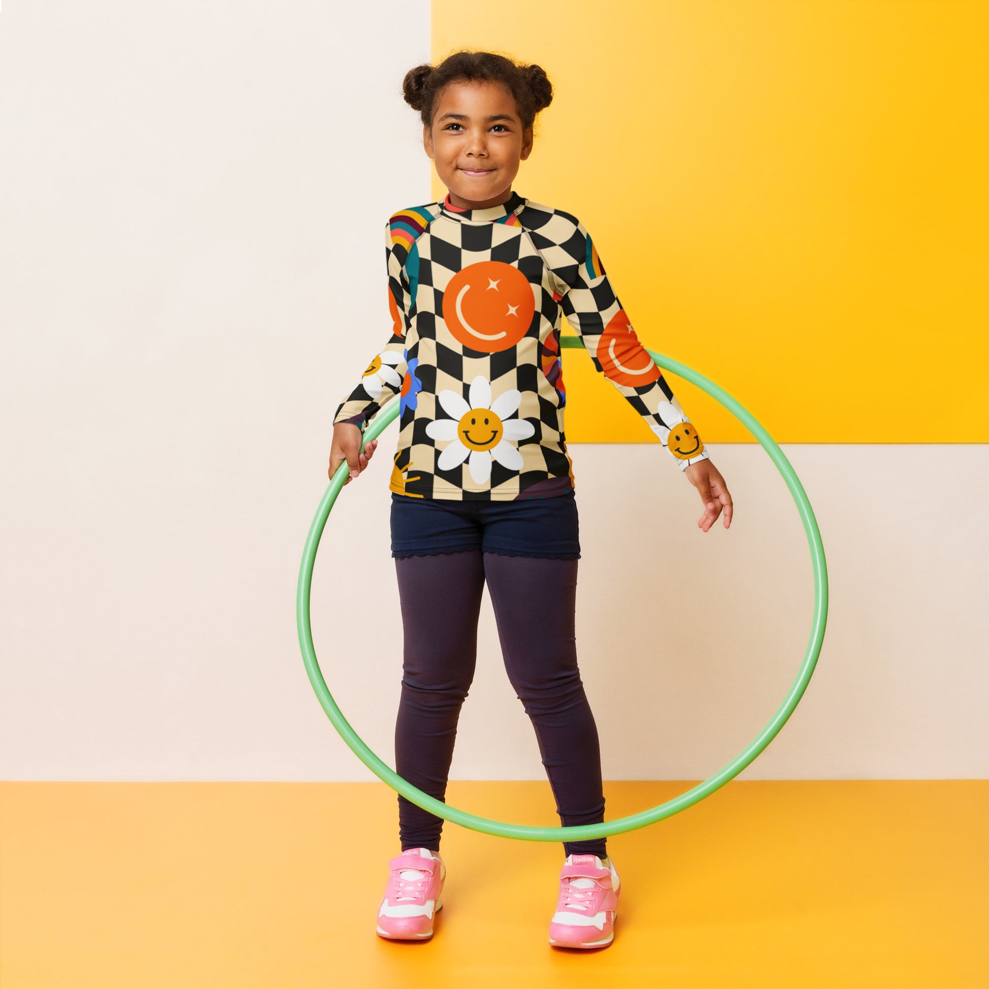 Tween girl hula hooping while wearing a retro wavy checkerboard girls rash guard with colorful smiley faces, rainbows, daisies, stars, and swirls. Long sleeve UPF 50+ swim shirt in sizes 2T–20 with coordinating family swimwear.