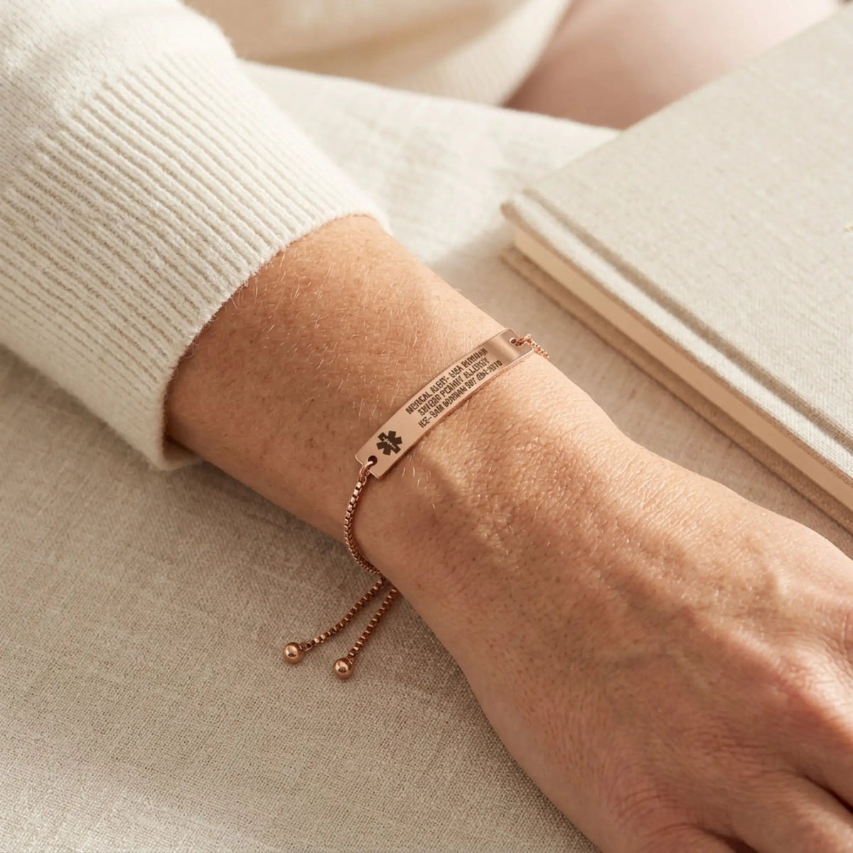 Rose gold personalized medical alert bracelet with a rectangular ID plate and adjustable sliding chain, worn on a woman's wrist with an ivory knit sweater. A soft neutral background and warm natural light highlight the bracelet's elegant finish and everyday wearable design.