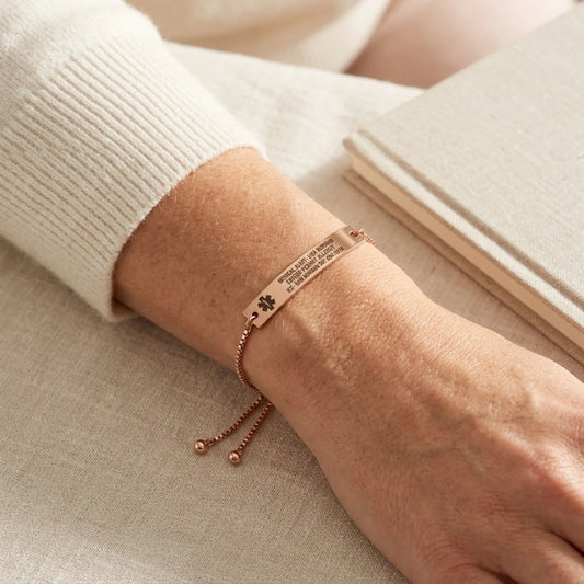Rose gold personalized medical alert bracelet with a rectangular ID plate and adjustable sliding chain, worn on a woman's wrist with an ivory knit sweater. A soft neutral background and warm natural light highlight the bracelet's elegant finish and everyday wearable design.